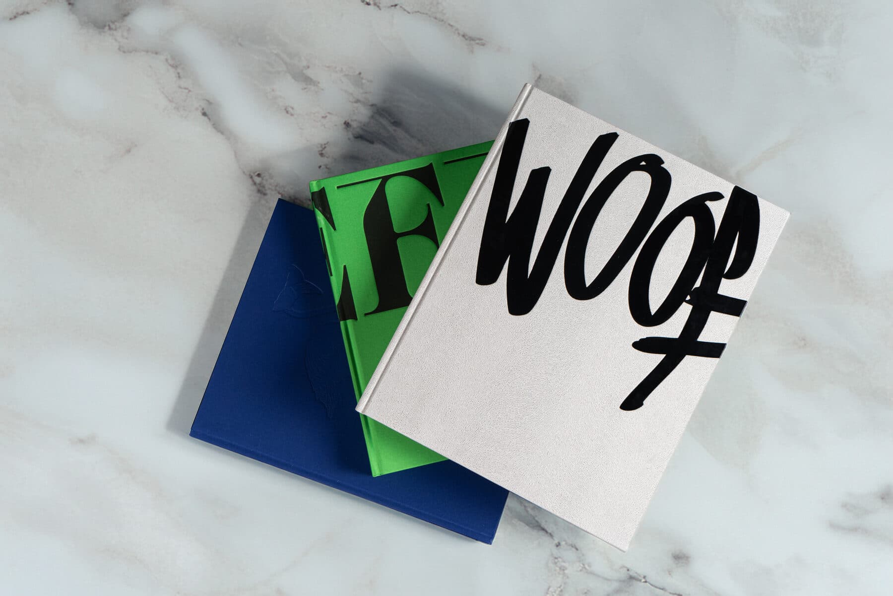 Top-down on marble: three editorial books stacked — royal blue, emerald green, and WOOF cream.